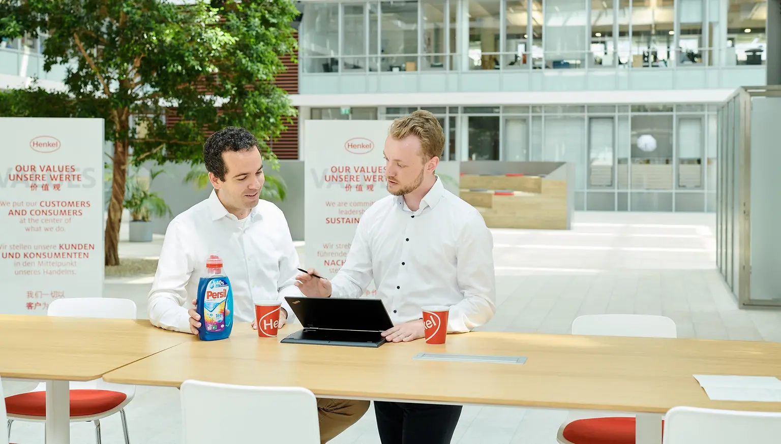 Dvaja muži stoja pri stole, na ktorom je položená nádoba Persil a laptop.